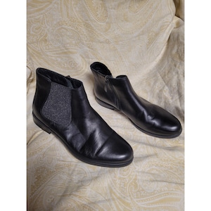 May include: A pair of black leather ankle boots featuring a smooth finish, a rounded toe, and a side zip closure. The boots include a dark grey elastic side panel with a subtle glitter texture, a low heel, and a black rubber outsole.