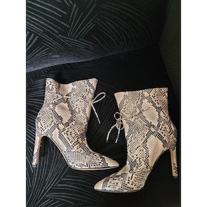 May include: A pair of ankle boots featuring a beige and black snakeskin pattern, a pointed toe, a thin stiletto heel, and a decorative lace-up detail at the top of the shaft. The boots are constructed with a textured material and a slim silhouette.