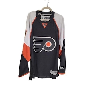 May include: Black, white, and orange hockey jersey with the Philadelphia Flyers logo on the front. The jersey has long sleeves, a V-neck, and the Reebok logo on the collar. The jersey is hanging on a hanger.