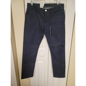 May include: Dark blue denim jeans featuring a classic five-pocket design, a silver-toned metal button closure at the waist, and a thin black drawstring cord hanging from the front. The waistband includes a visible Armani Exchange brand label.