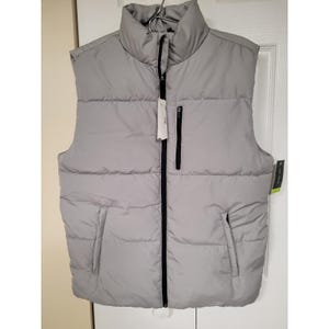 May include: A light gray puffer vest with a high collar and a black zipper closure. The vest has two side pockets and a small chest pocket with a black zipper. The vest is hanging on a metal hanger.