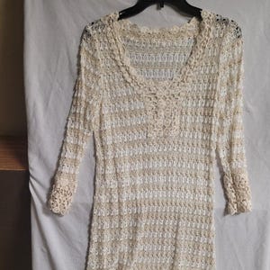 May include: A cream-colored, long-sleeved crochet top with a V-neckline. The top features a delicate, open-weave pattern with a scalloped neckline and cuffs. The crochet design includes a series of small, decorative buttons down the center.