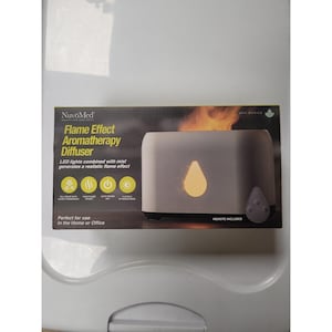Puede incluir: Caja de un difusor de aromaterapia NuvoMed con efecto de llama. La caja gris muestra un difusor con una luz en forma de llama. El texto de la caja dice: "Las luces LED combinadas con la niebla generan un efecto de llama realista."