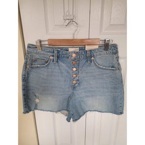 Könnte beinhalten: Hellblaue Jeansshorts mit Knopfleiste und rohem Saum. Die Shorts haben eine hohe Taille und ein Distressed-Detail auf der Vorderseite. Sie hängen an einem Holzbügel.