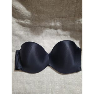 May include: A strapless black bra with a smooth, satin-like finish. The bra has a curved neckline and is designed to provide support without straps. The fabric appears to be a solid, dark color, and the bra is laid flat on a textured surface.