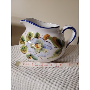 Op de afbeelding: Witte keramische kan met een blauwe rand en handvat, versierd met handgeschilderde bloemmotieven in blauw, groen en oranje. De kan is ongeveer 15 cm hoog en is geschikt voor het serveren van dranken.