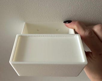 Puede incluir: Un contenedor de almacenamiento de plástico blanco y rectangular con tapa. El contenedor está sostenido por una mano con esmalte de uñas oscuro. El contenedor está montado sobre una superficie blanca.