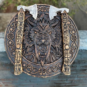 May include: A wooden wall hanging featuring a carved wolf's head and intricate knotwork. Two decorative axes with silver blades and wooden handles are mounted on either side of the wolf's head. The piece is stained in shades of brown.