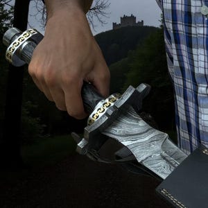 May include: A Damascus steel dagger with a patterned blade and a decorative handle with gold-colored chain accents. The dagger is held in a hand, with a castle in the background.