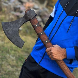 May include: A handcrafted axe with a dark, weathered blade and a wooden handle wrapped in brown leather. The handle is adorned with brass studs. The axe is held at an angle, showcasing its design and craftsmanship.