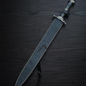 May include: A black sword with a leather sheath, resting on a dark wooden surface. The sword features a detailed handle and guard, with a black leather strap. The sheath is stitched with white thread.