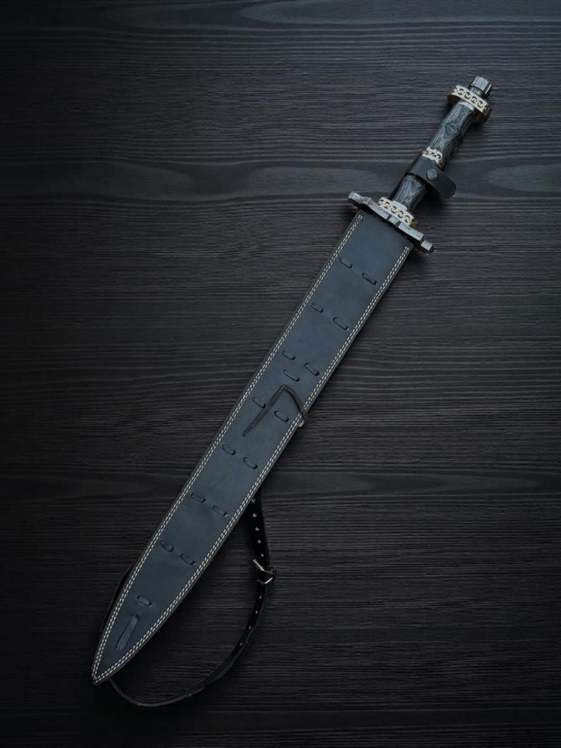 May include: A black sword with a leather sheath, resting on a dark wooden surface. The sword features a detailed handle and guard, with a black leather strap. The sheath is stitched with white thread.