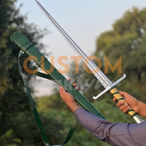 May include: A long, silver sword with a gold and green handle is held next to a green sheath. The sword has a crossguard and a long, sharp blade. The sheath has a strap for carrying. The word "CUSTOM" is partially visible.
