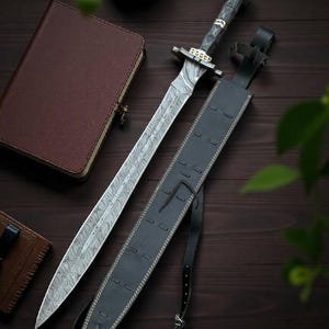 May include: A Damascus steel sword with a patterned blade and a dark wooden handle, resting beside a black leather sheath. The sword features gold-colored accents and is displayed on a dark wooden surface, alongside a leather-bound journal.
