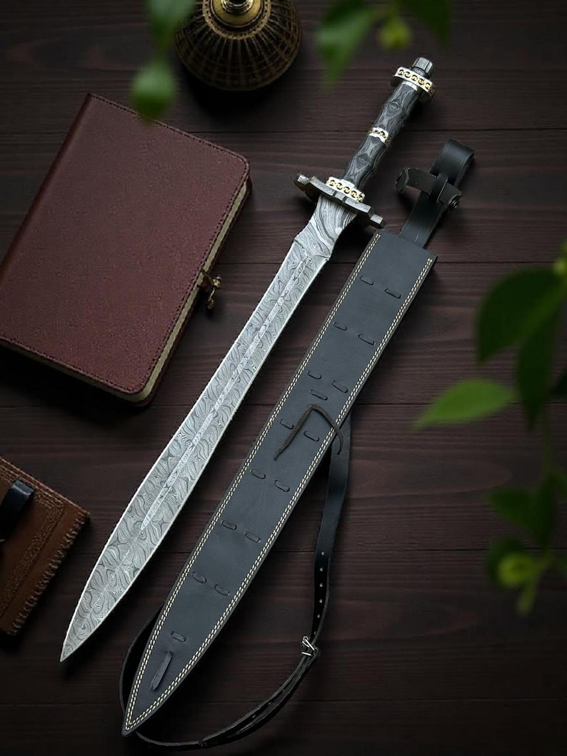May include: A Damascus steel sword with a patterned blade and a dark wooden handle, resting beside a black leather sheath. The sword features gold-colored accents and is displayed on a dark wooden surface, alongside a leather-bound journal.