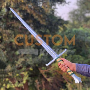 May include: A long, silver sword with a double-edged blade and a cross-shaped hilt. The handle is wrapped in green and gold material. The text "CUSTOM KNIVES" is visible on the blade.