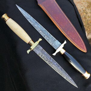 Könnte beinhalten: Zwei verzierte Dolche mit gemusterten Klingen und dekorativen Griffen. Ein Dolch hat einen hellfarbigen Griff mit goldenen Akzenten, der andere einen schwarzen Griff. Eine braune Lederscheide mit dekorativen Nähten ist ebenfalls vorhanden.