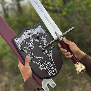 May include: A longsword with a silver blade featuring engraved text, a maroon handle, and a gold lion head pommel. The sword is paired with a black shield-shaped plaque displaying a white lion graphic and a maroon scabbard with a silver tip.