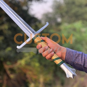 May include: A long, silver sword with a polished blade and a decorative handle. The handle features a gold grip with green accents. The sword's design includes a crossguard and a pointed pommel. The sword is held in a person's hand.