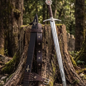 May include: A steel sword with a leather-wrapped hilt and a circular pommel featuring a red cross emblem. The blade is engraved with text. Beside it is a dark brown leather scabbard and belt adorned with small gold-colored cross accents.