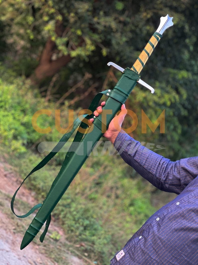 May include: A long sword with a silver blade and a wooden handle wrapped in gold and green. The sword is in a green sheath with a shoulder strap. The sword is held by a person.