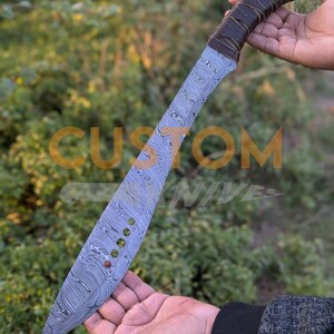 以下が含まれることがあります： 模様のある刃と濃い茶色の革で包まれたハンドルが付いた、大きなダマスカス鋼のナイフ。 刃には、一連の小さな装飾的なリベットが付いています。 背景には「CUSTOM KNIVES」の文字が見えます。