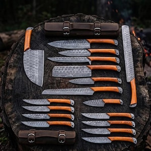 May include: A collection of various kitchen knives featuring patterned Damascus steel blades and light brown wooden handles. The set includes a large cleaver, a long rectangular blade, and multiple utility knives, accompanied by two brown leather cases.