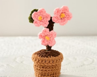 May include: A handcrafted crochet plant with three pink flowers, yellow centers, and a green leaf. The flowers are attached to a brown stem, planted in a brown crochet pot. The pot is sitting on a white surface.