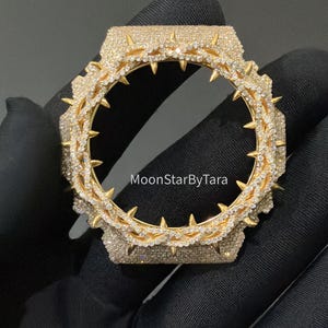 May include: A gold-toned watch bezel featuring a crown of thorns design encrusted with small clear stones. The circular frame has sharp, protruding spikes and a pavé-set surface. The text MoonStarByTara is printed in the center of the bezel.