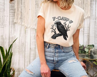 May include: Cream-colored t-shirt with a graphic of a black crow perched on a branch, set against a full moon. The text "F-CAW-F" is arched above the crow. The shirt is paired with light blue denim jeans.