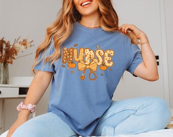 May include: A blue t-shirt with the word "NURSE" in a fall-themed design. The letters are decorated with floral patterns and a gingham pattern. The design includes pumpkins, leaves, and a stethoscope.