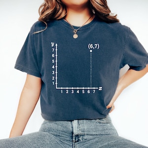 May include: Navy blue t-shirt featuring a white coordinate plane graphic with the point (6,7) plotted. The x-axis and y-axis are labeled. The shirt is made of a soft material and has short sleeves.