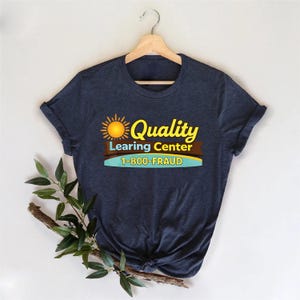 May include: Navy blue t-shirt with the text "Quality Learing Center 1-800-FRAUD" in yellow, brown, and blue. The design includes a sun graphic. The shirt is displayed on a wooden hanger.