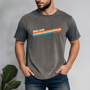 May include: Dark grey t-shirt with the text "REBEL SCUM" above a retro-style design with the words "SINCE 1977". The design features horizontal stripes in orange, yellow, blue, and teal.
