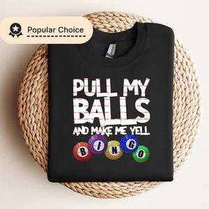 Könnte beinhalten: Schwarzes Sweatshirt mit dem weißen Text "PULL MY BALLS AND MAKE ME YELL". Darunter befinden sich Bingokugeln mit den Buchstaben B, I, N, G und O. Das Sweatshirt liegt auf einer gewebten, kreisförmigen Oberfläche.