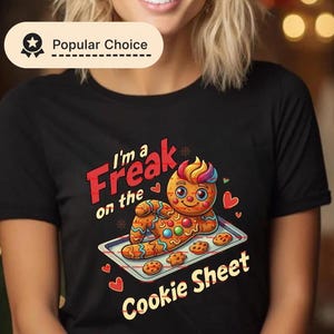 May include: Black t-shirt featuring a colorful gingerbread man design on a cookie sheet. The text reads "I'm a Freak on the Cookie Sheet" in red and white. The design includes hearts and stars.