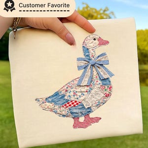 May include: A cream-colored square pillow featuring a patchwork goose design. The goose has a blue gingham bow, red and white checkered feet, and a beak. The pillow is held by a person.