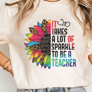 Puede incluir: Camiseta color crema con un diseño de girasol colorido. El girasol tiene pétalos de arcoíris y un acento de estampado de leopardo. El texto dice: "IT TAKES A LOT OF SPARKLE TO BE A TEACHER."