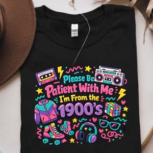 Könnte beinhalten: Schwarzes T-Shirt mit einem farbenfrohen Retro-Design. Die Grafik zeigt einen Ghettoblaster, eine Kassette, Rollschuhe und den Text "Please Be Patient With Me I'm From The 1900's". Weitere Elemente sind ein Rubik's Cube, Sonnenbrille und Kopfhörer.