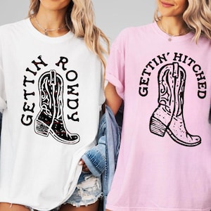 Könnte beinhalten: Zwei T-Shirts, eines weiß und eines rosa, jeweils mit einer Cowboy-Stiefel-Grafik. Das weiße Shirt trägt den Schriftzug "Gettin' Rowdy" und das rosa Shirt "Gettin' Hitched" in schwarzer Schrift. Die Stiefel sind schwarz mit weißen und roten Akzenten.