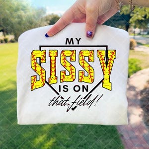 以下が含まれることがあります： 「MY SISSY IS ON that field!」の文字が黄色と赤のソフトボール風の文字でプリントされた白いTシャツ。デザインには、黒い野球のダイヤモンドの輪郭が含まれています。