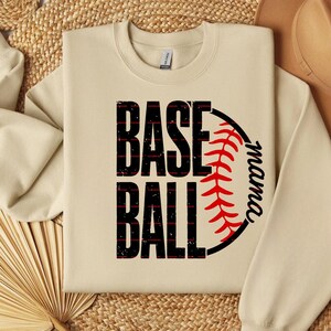 Op de afbeelding: Een beige sweater met ronde hals voorzien van een grafische print met de zwarte, verweerde tekst BASE BALL verticaal gestapeld. Rechts een gedeeltelijke honkbalafbeelding met rode stiksels en het zwarte cursieve woord mama langs de rand.