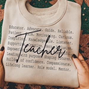 Puede incluir: Una sudadera beige con la palabra "Teacher" en cursiva. La sudadera tiene cuello redondo acanalado y enumera atributos positivos de un maestro en fuente negra.