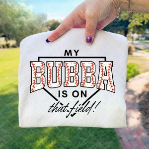 May include: A white t-shirt with the text "MY BUBBA IS ON that field!" in black and red. The word "BUBBA" is designed to look like a baseball. The shirt is held up by a hand with a blue fingernail.