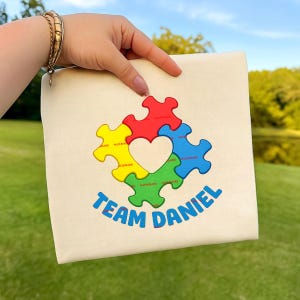 Op de afbeelding: Een crèmekleurige draagtas met een kleurrijk puzzelstukontwerp en een hartvormige uitsparing. De woorden "TEAM DANIEL" staan eronder in het blauw gedrukt. De tas wordt vastgehouden tegen een groene grasachtergrond.