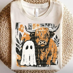 May include: Cream-colored t-shirt featuring a distressed graphic of a Highland cow and a ghost. The cow is in shades of orange and black, with a ghost to the left. Floral and leaf accents surround the cow and ghost.