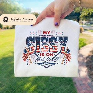 May include: White t-shirt with the text "MY SISSY IS ON that field!" in red, white, and blue, with American flags and fireworks graphics. The shirt is a popular choice.