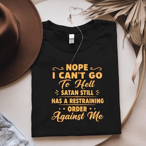 May include: A black short-sleeved t-shirt featuring yellow text that reads: NOPE I CAN'T GO To Hell SATAN STILL HAS A RESTRAINING ORDER Against Me. The shirt has a crew neckline and a white brand label visible at the inner collar area.