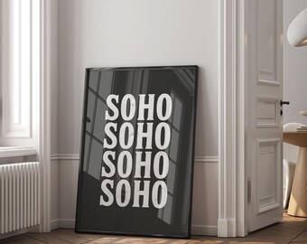 May include: A framed black and white art print leaning against a wall. The print features the word "SOHO" stacked four times in a bold, white font. The artwork is displayed in a room with wooden flooring and a white door.