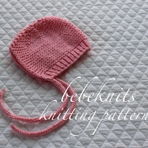 May include: A pink knitted baby bonnet with ties. The bonnet is on a white quilted fabric background. The text "bebeknits knitting pattern" is written in cursive below the bonnet.